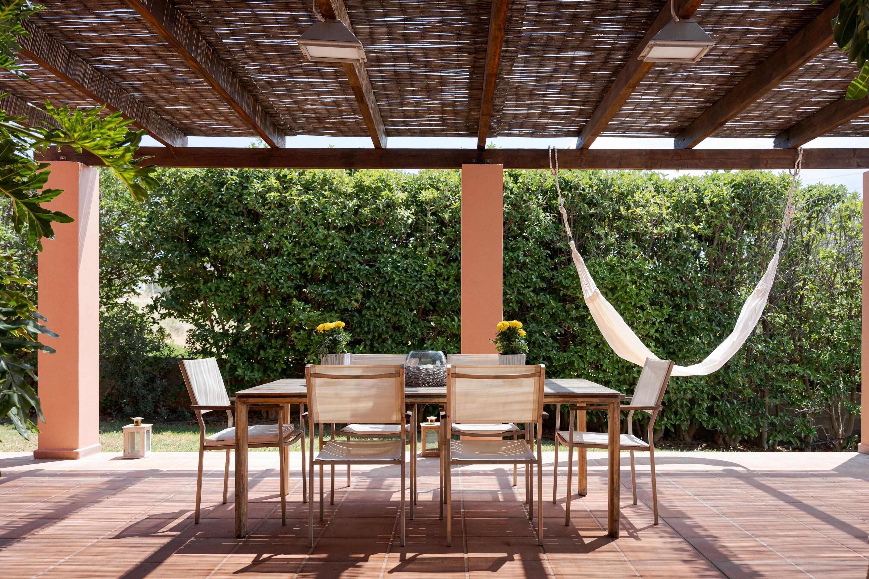 An outdoor patio with a wooden table and chairs, a hanging hammock, and a backdrop of dense foliage.