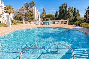 A swimming pool with a tiled edge, surrounded by a white fence, palm trees, and a building with balconies in the background. - Luxury Family Riviera Del Mar (Mijas)