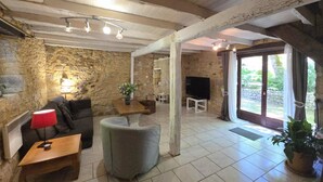 A rustic interior with stone walls, a wooden ceiling, a sofa, a small table with a lamp, a TV, and a large window.