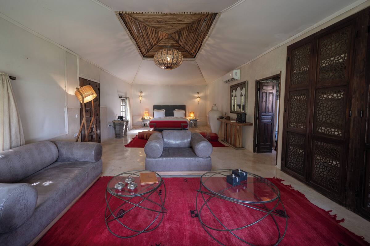 A spacious living room with a large sofa, two glass coffee tables, a red rug, a bed with a grey headboard, and a decorative ceiling.