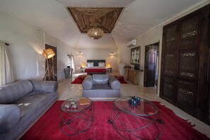 Un salon spacieux avec un grand canapé, deux tables basses en verre, un tapis rouge, un lit avec une tête de lit grise et un plafond décoratif.