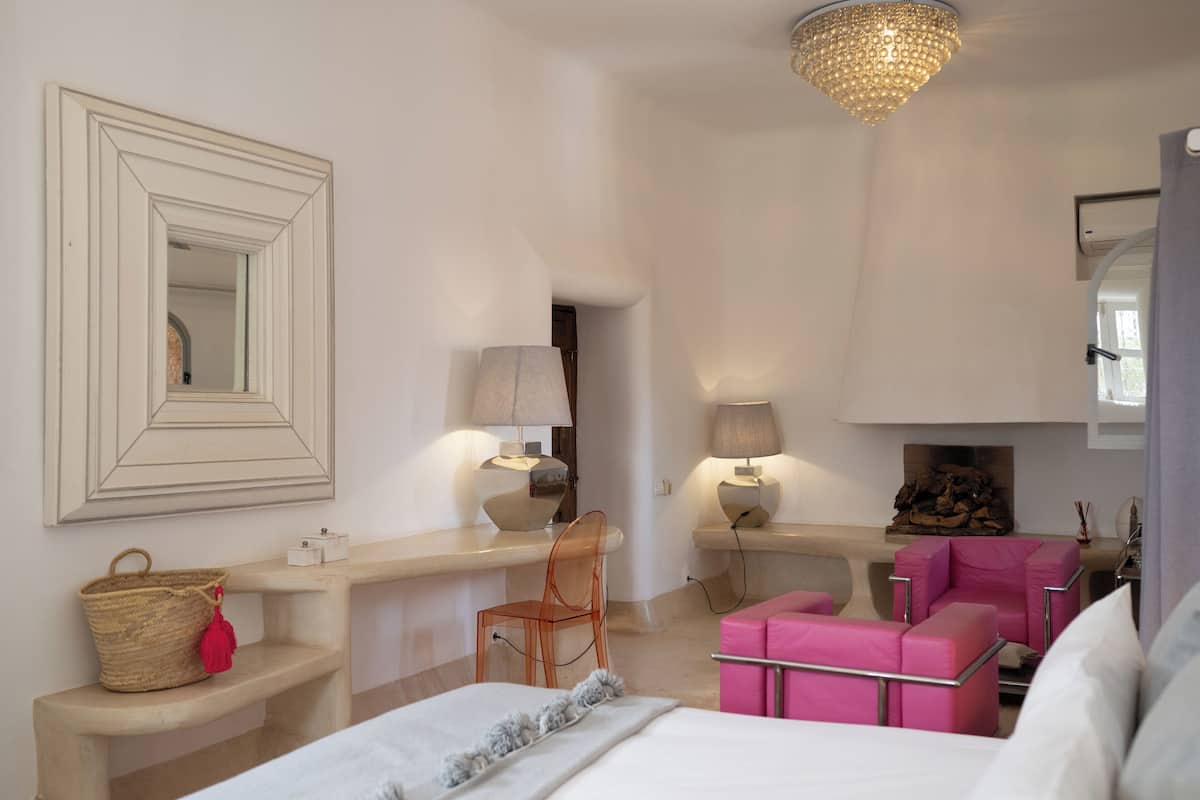 A bedroom with a bed, a desk, a chair, a pink armchair, a fireplace, and a mirror on the wall.