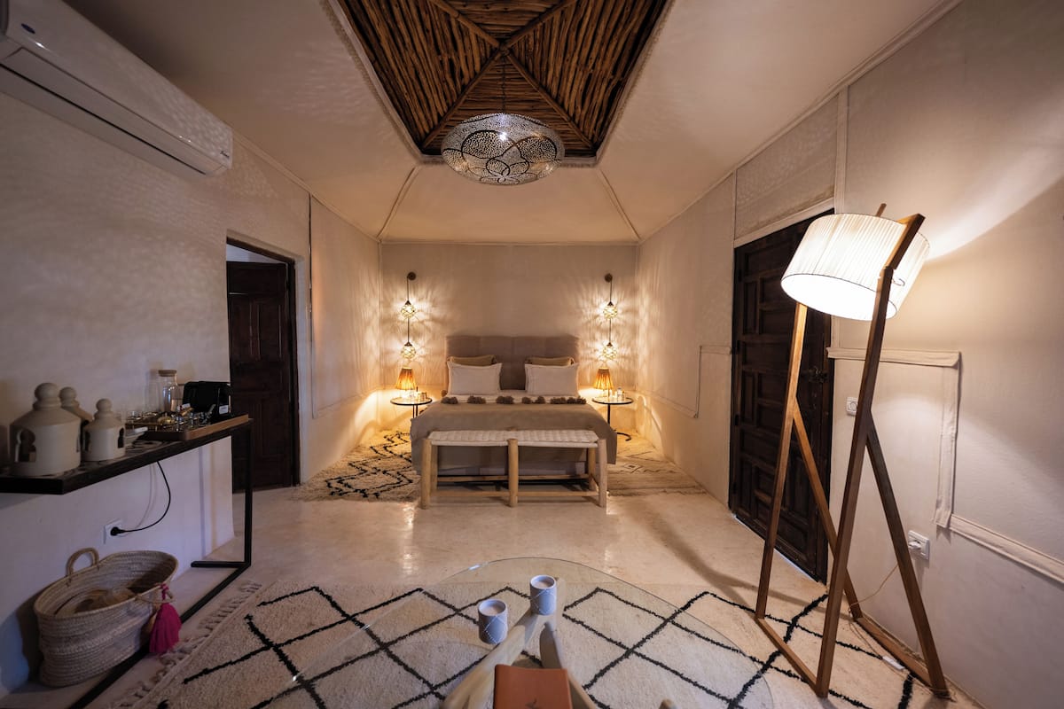 A bedroom with a bed, bedside lamps, a bedside table, a floor lamp, a wall-mounted shelf, and a patterned rug.