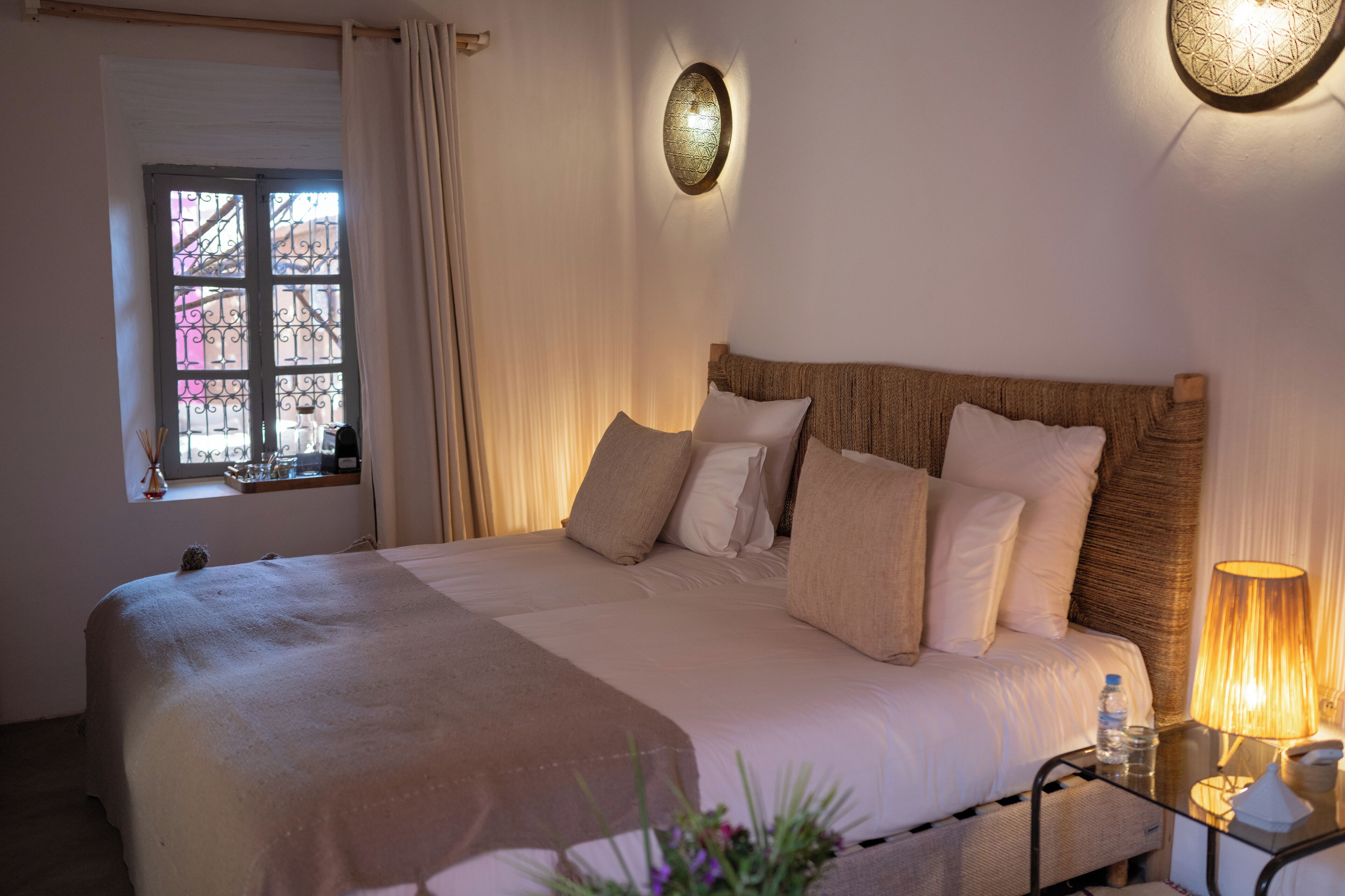 A neatly made bed with a headboard, a bedside table with a lamp, and a window with curtains.