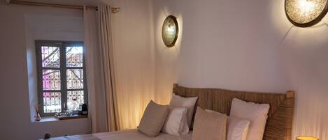 A neatly made bed with a headboard, a bedside table with a lamp, and a window with curtains.
