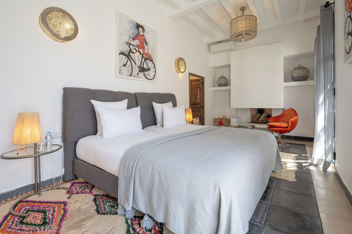 A modern bedroom with a large bed, a side table with a lamp, a colorful rug, and a bathroom visible in the background.