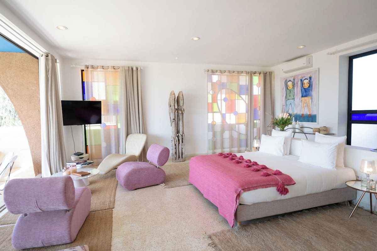 A modern hotel room with a large bed, a flat-screen TV, a seating area with chairs, and a large window with curtains.