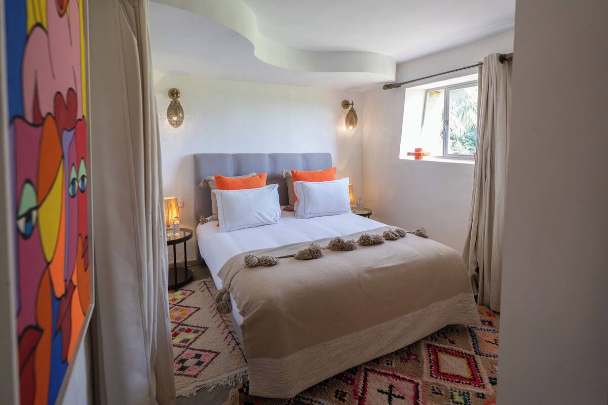 A bedroom with a bed, two bedside tables, a window, and a wall-mounted light fixture.