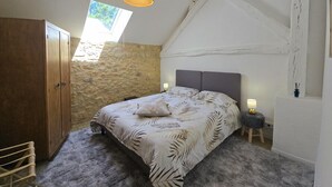 A bedroom with a sloped ceiling, a bed with a leaf-patterned bedspread, a wooden wardrobe, and a stone wall. - Charming Cottage with Pool and Private Parking in Périgord Noir (Berbiguières)
