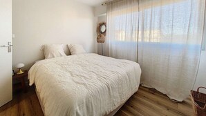 A bedroom with a large bed, a bedside table with a lamp, a mirror, and a window with sheer curtains. - Ideal Beach T2 Cabin with Parking, Pets Allowed (Cucq-Stella Plage)