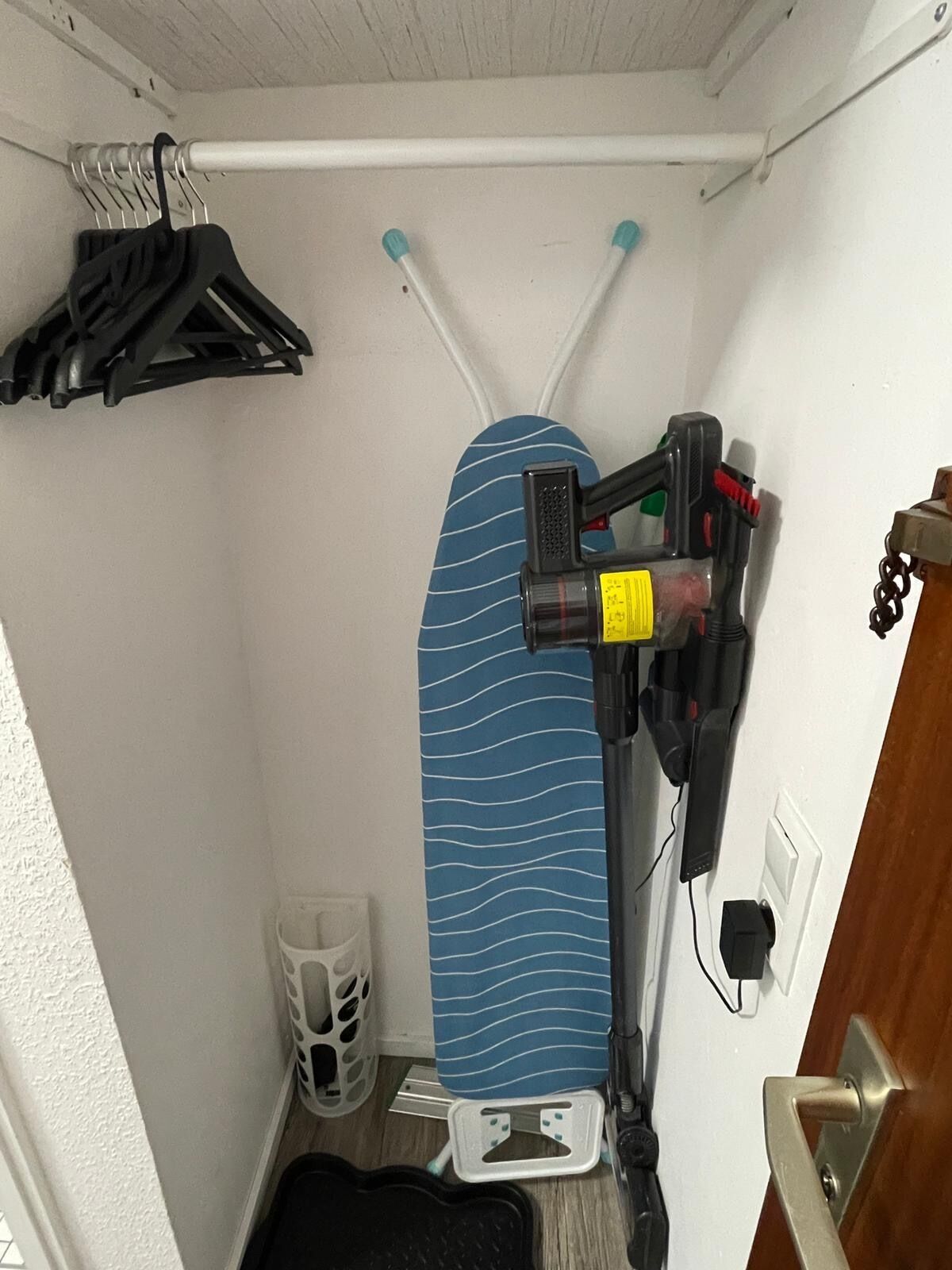 Ein kleines Zimmer mit einem Bett, einer kleinen Ablage, einer Kleiderstange mit Kleidung und einer Tür mit Holzrahmen.