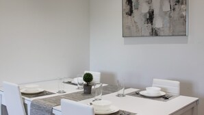 Ein Essbereich mit einem weißen Tisch, gedeckt für vier Personen, inklusive Teller, Gläser und Besteck. Auf dem Tisch steht ein kleiner Topfpflanze, und darüber an der Wand hängt ein großes abstraktes Gemälde.