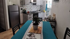 Eine kleine Küche mit Kühlschrank, Spüle und Arbeitsfläche. Ein Esstisch mit blauer Tischdecke und floralem Untersetzer.