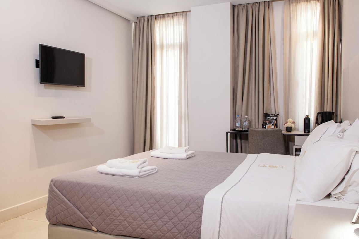 Une chambre d’hôtel avec un lit, un téléviseur à écran plat fixé au mur, une petite table avec une machine à café et une fenêtre avec des rideaux.