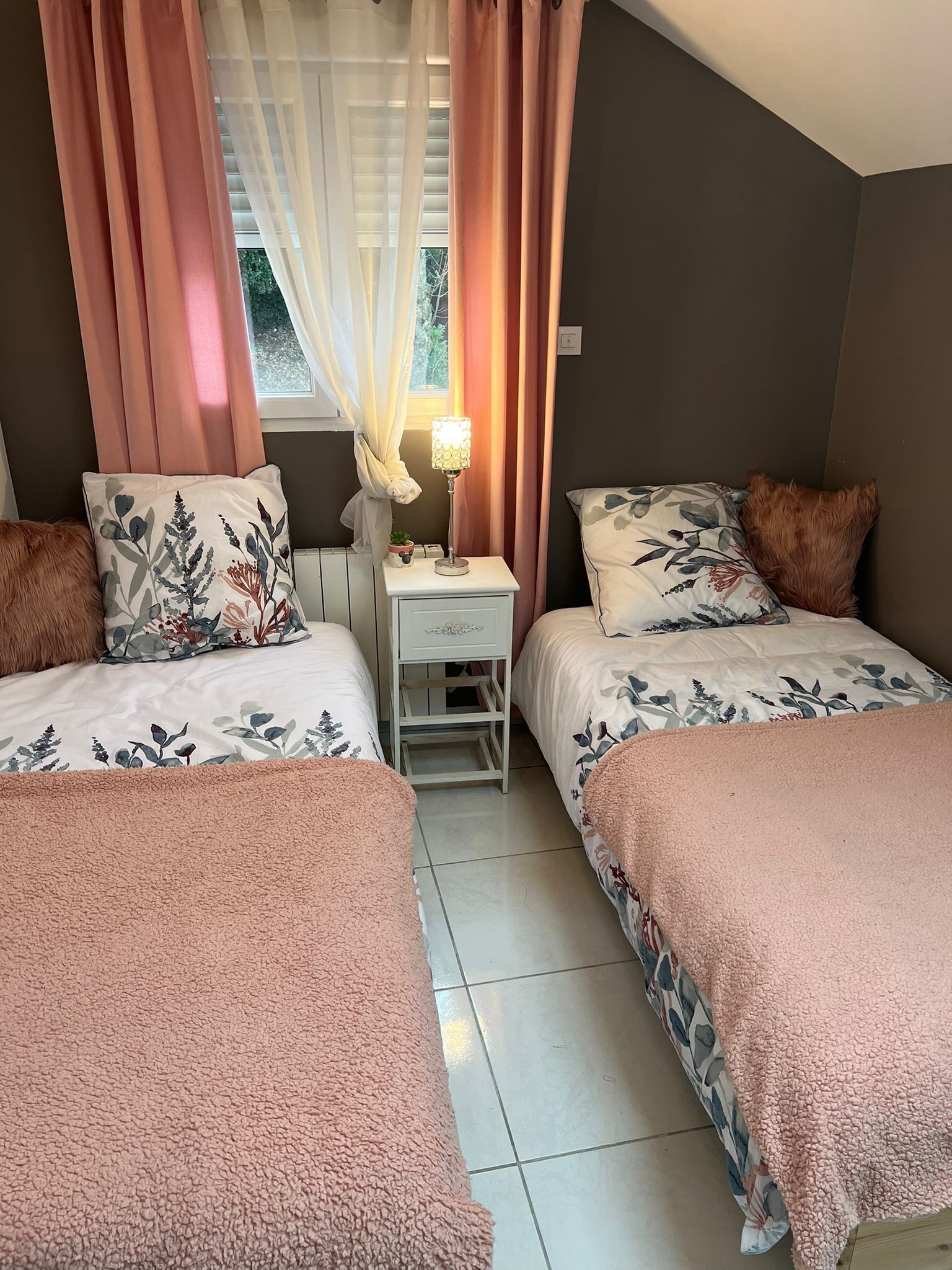 A bedroom with two beds, a nightstand with a lamp, and a window with sheer curtains.