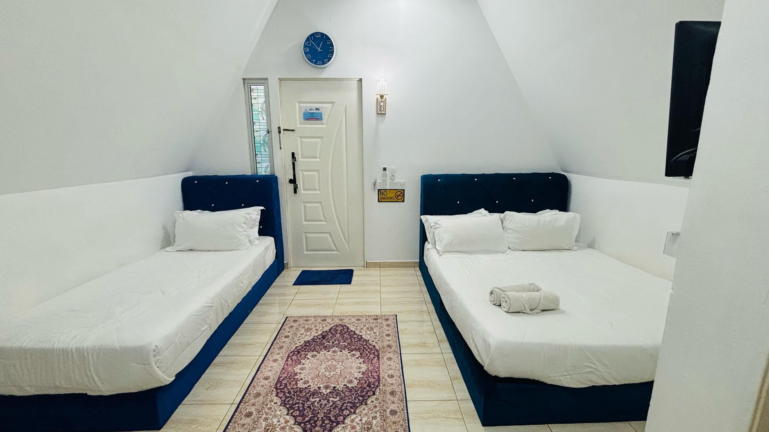 A room with two single beds, a door with a smoke alarm, a clock, and a small rug.