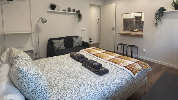 A modern bedroom with a large bed, a desk, and a kitchenette in the background.