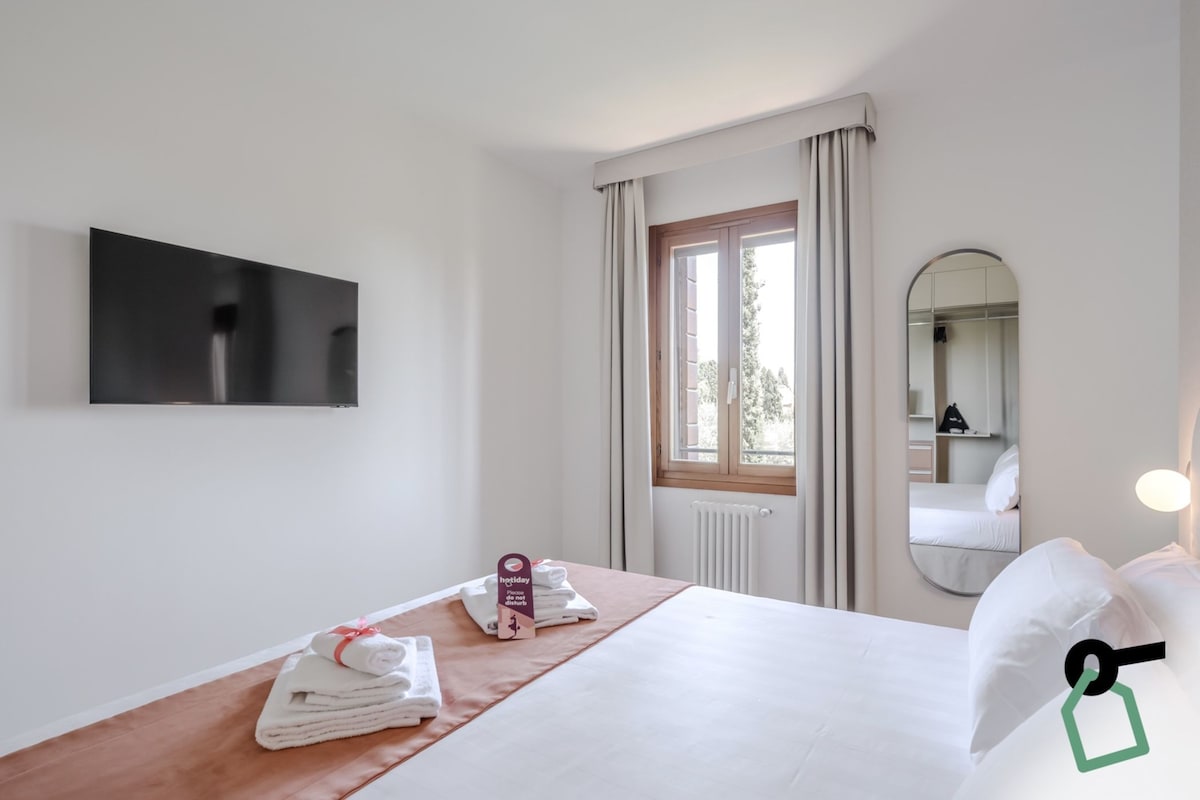 A hotel room with a bed, a TV mounted on the wall, a window with curtains, a bedside table with towels, and a mirror.