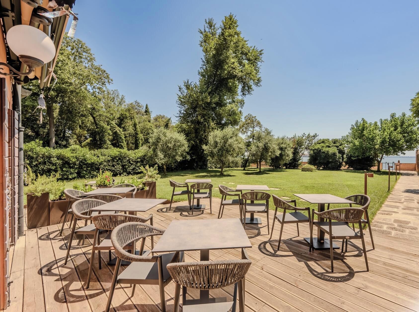 Een terras buiten met tafels en stoelen, omringd door groen en bomen.