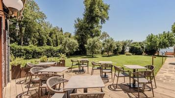 Een terras buiten met tafels en stoelen, omringd door groen en bomen.