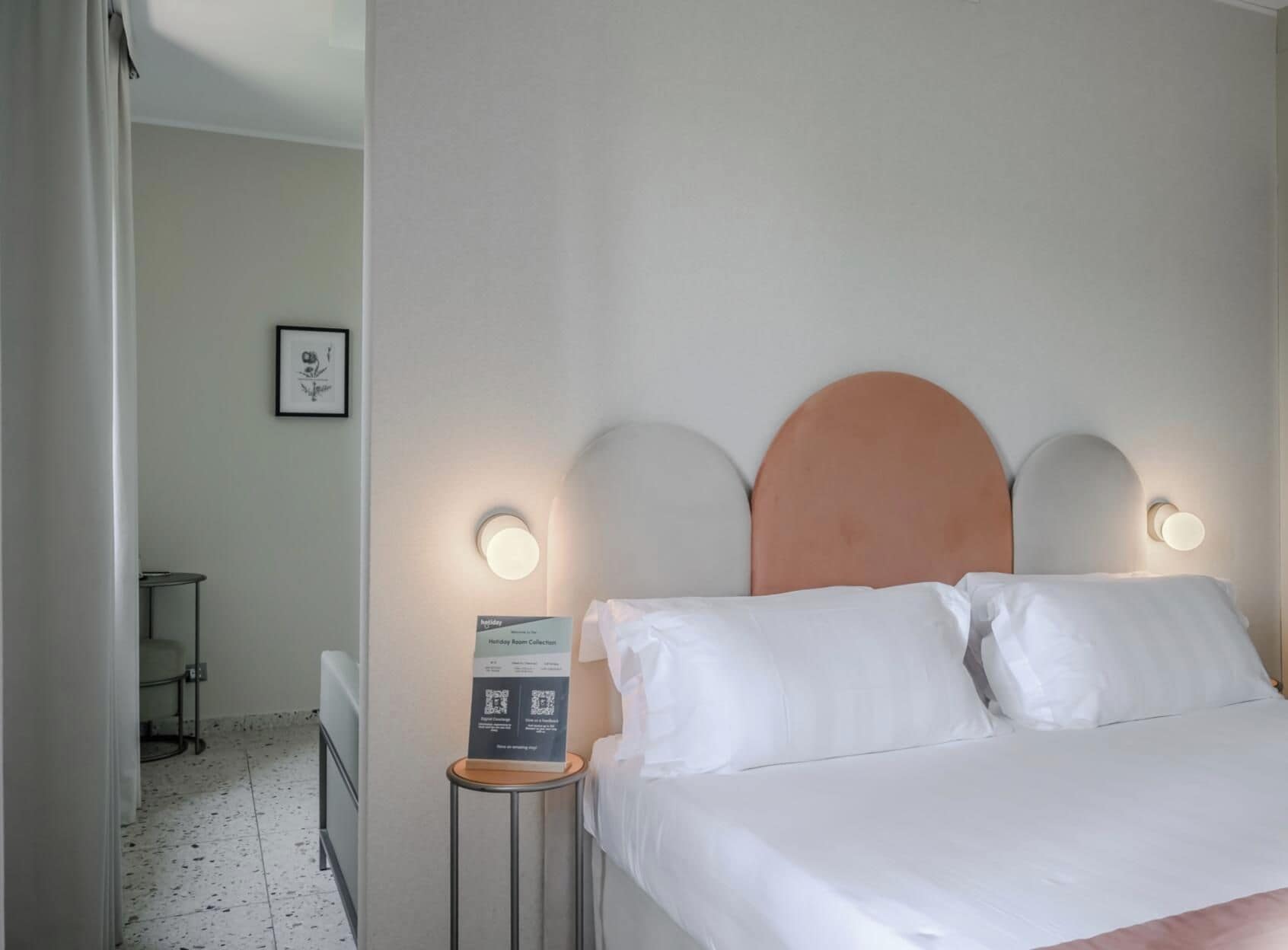 A hotel room with a bed, two bedside lamps, a small table, and a framed picture on the wall.