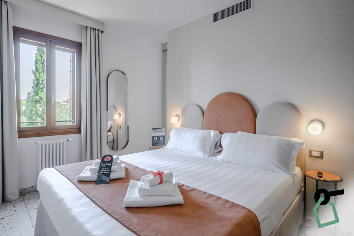 A hotel room with a bed, a bedside table, a mirror, and a window with curtains.