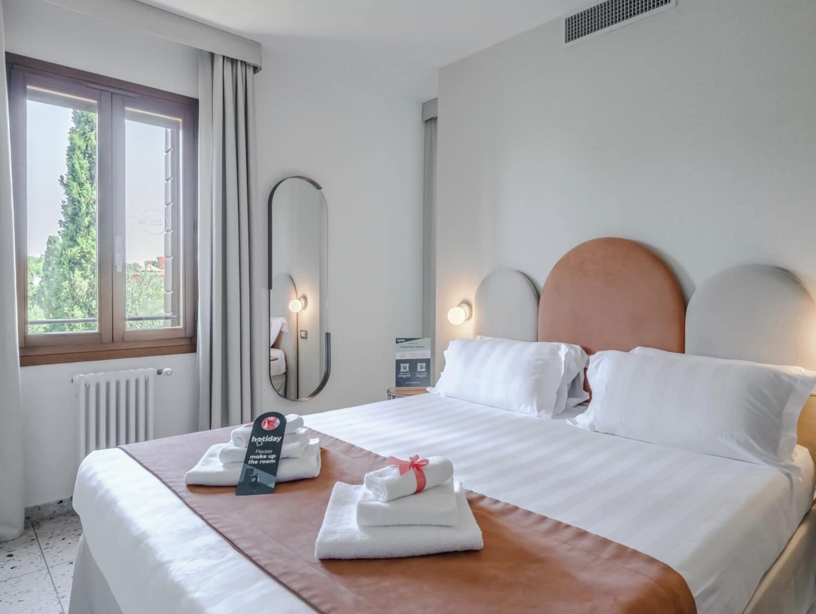 A hotel room with a bed, a large mirror, a window with curtains, and a sign indicating the room is ready for guests.