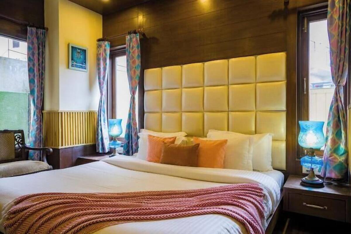 A hotel room with a large bed, a padded headboard, a bedside table with a lamp, and curtains.