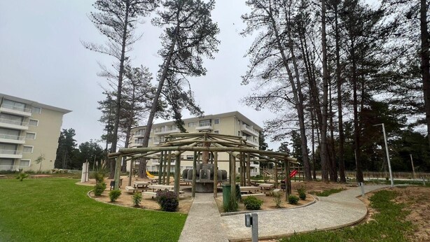 A park with a playground, picnic tables, and a building in the background. - Departamento acojedor en Parque mar4 Piso 1 (El Tabo)