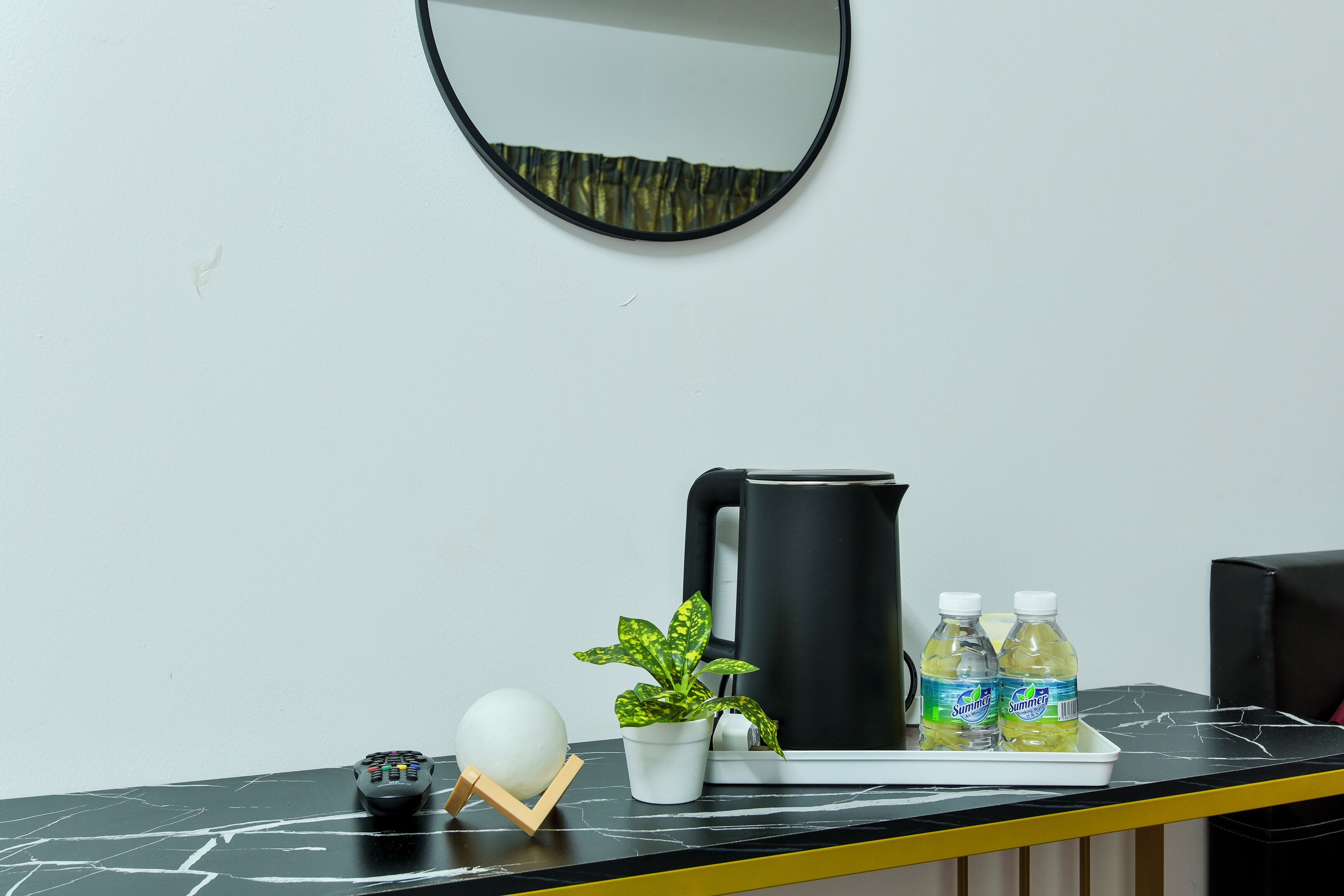A hotel reception desk with a black marble countertop, a white vase with a green plant, a black coffee maker, two bottles of water, a remote control, and a spherical object.
