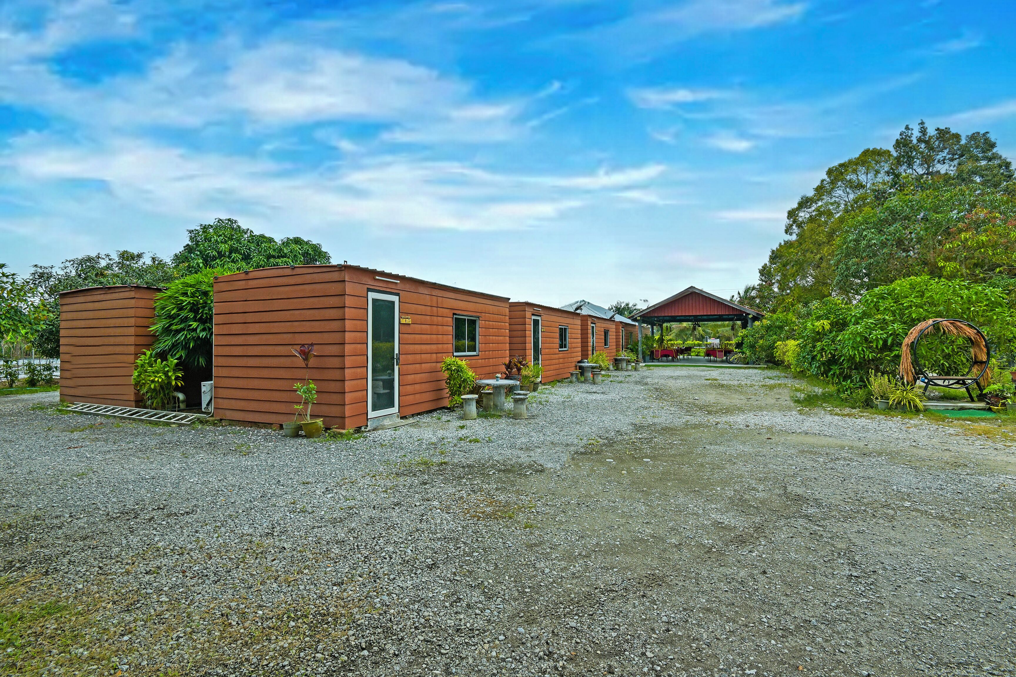 A row of brown buildings with a gravel driveway, surrounded by greenery and a clear sky.