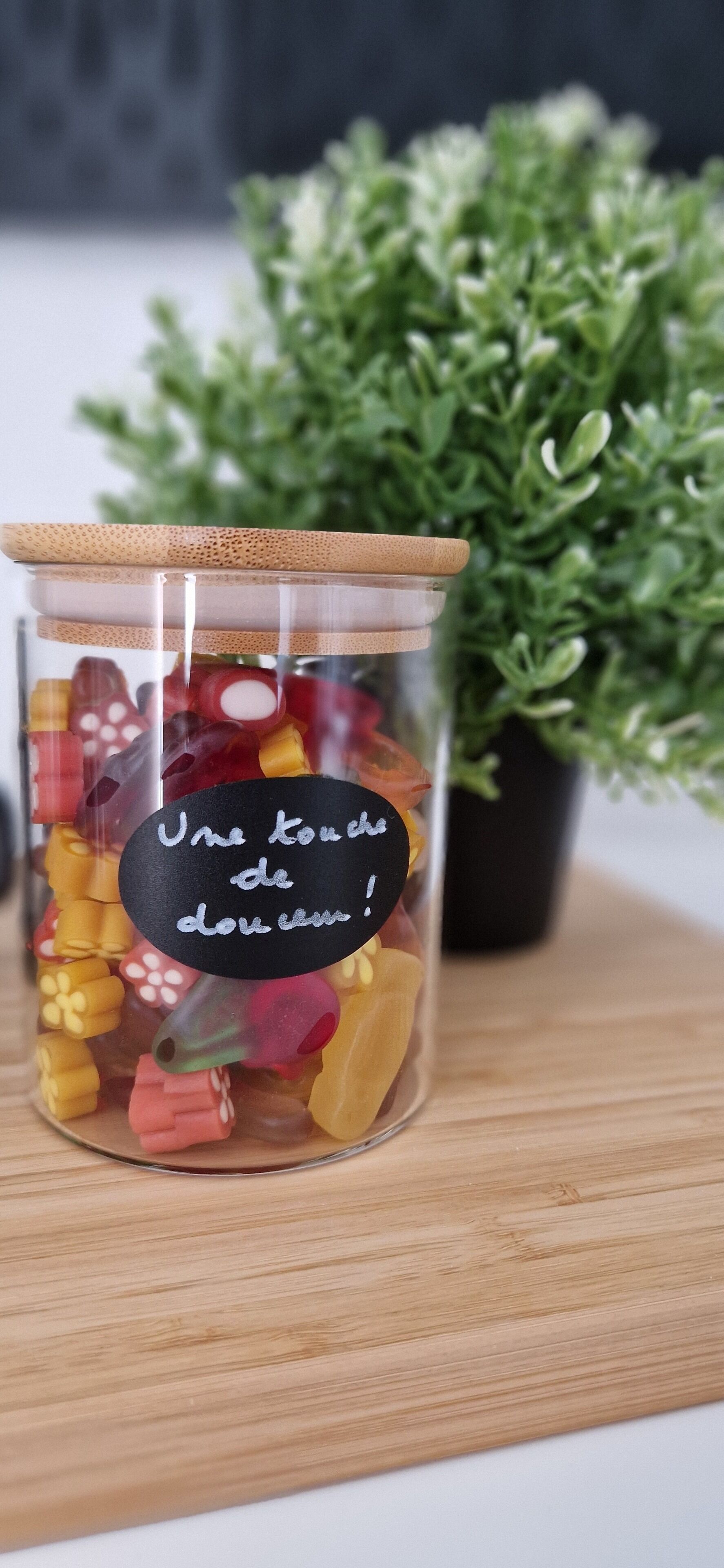 A glass jar filled with colorful gummy candies, with a label that reads "Une touche de douceur!" on a wooden surface with a potted plant in the background.
