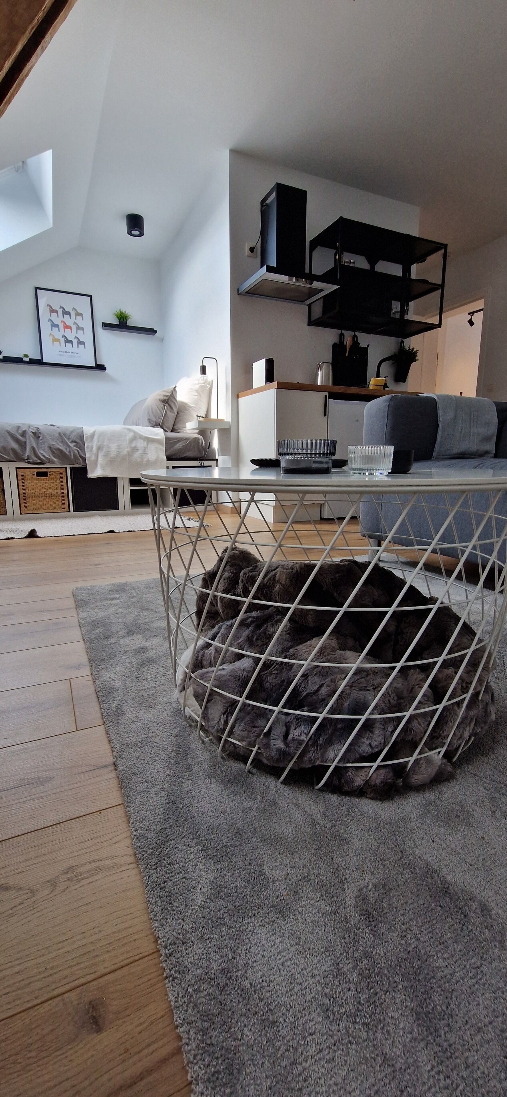 Uma sala de estar acolhedora com um sofá cinzento, uma mesa redonda pequena com abajur e uma caminha para animal de estimação com estrutura de arame.