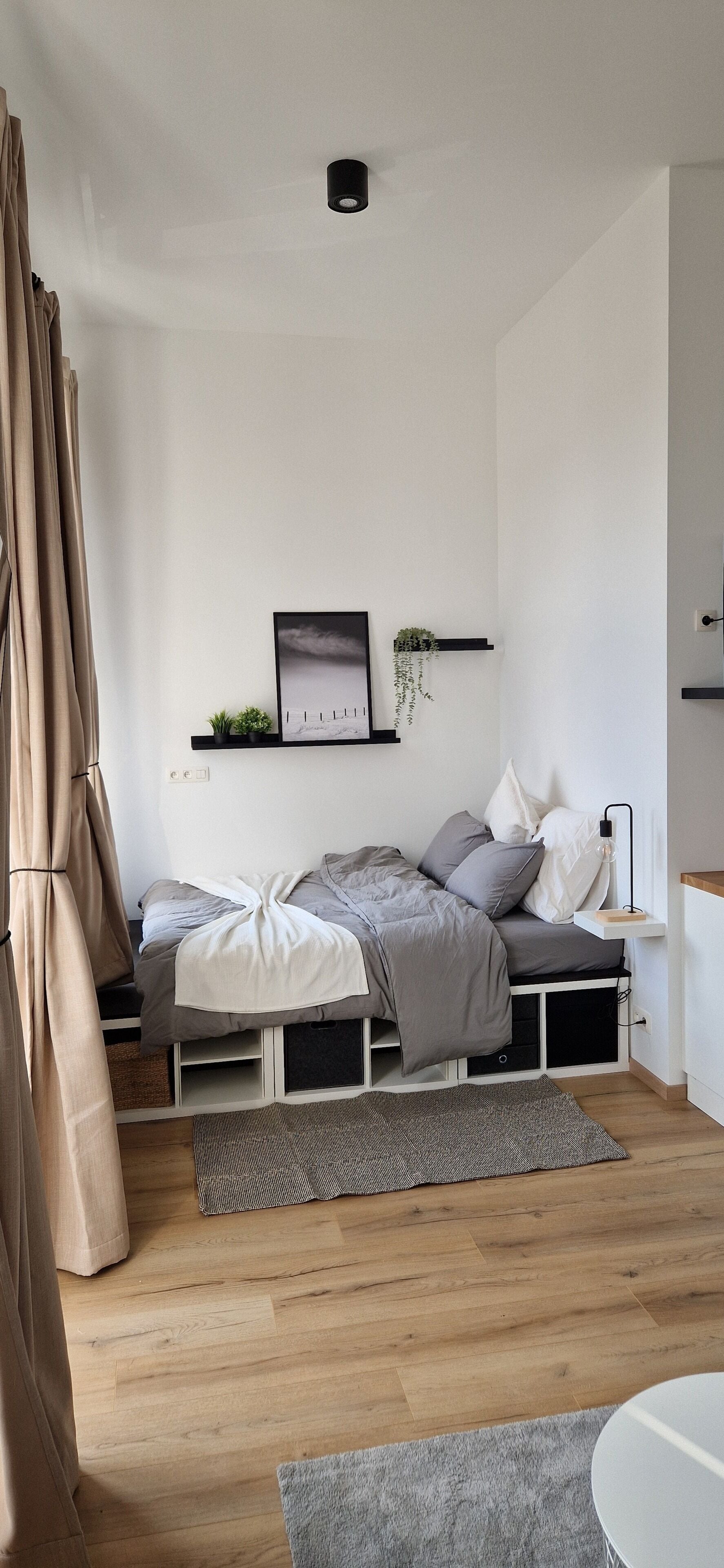 Un dormitorio moderno con cama, mesitas de noche, un pequeño estante con plantas y un cuadro en la pared.