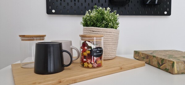 Una encimera de cocina con una taza negra, un tarro de cristal con caramelos de colores, una taza blanca y una cesta de mimbre con plantas verdes.