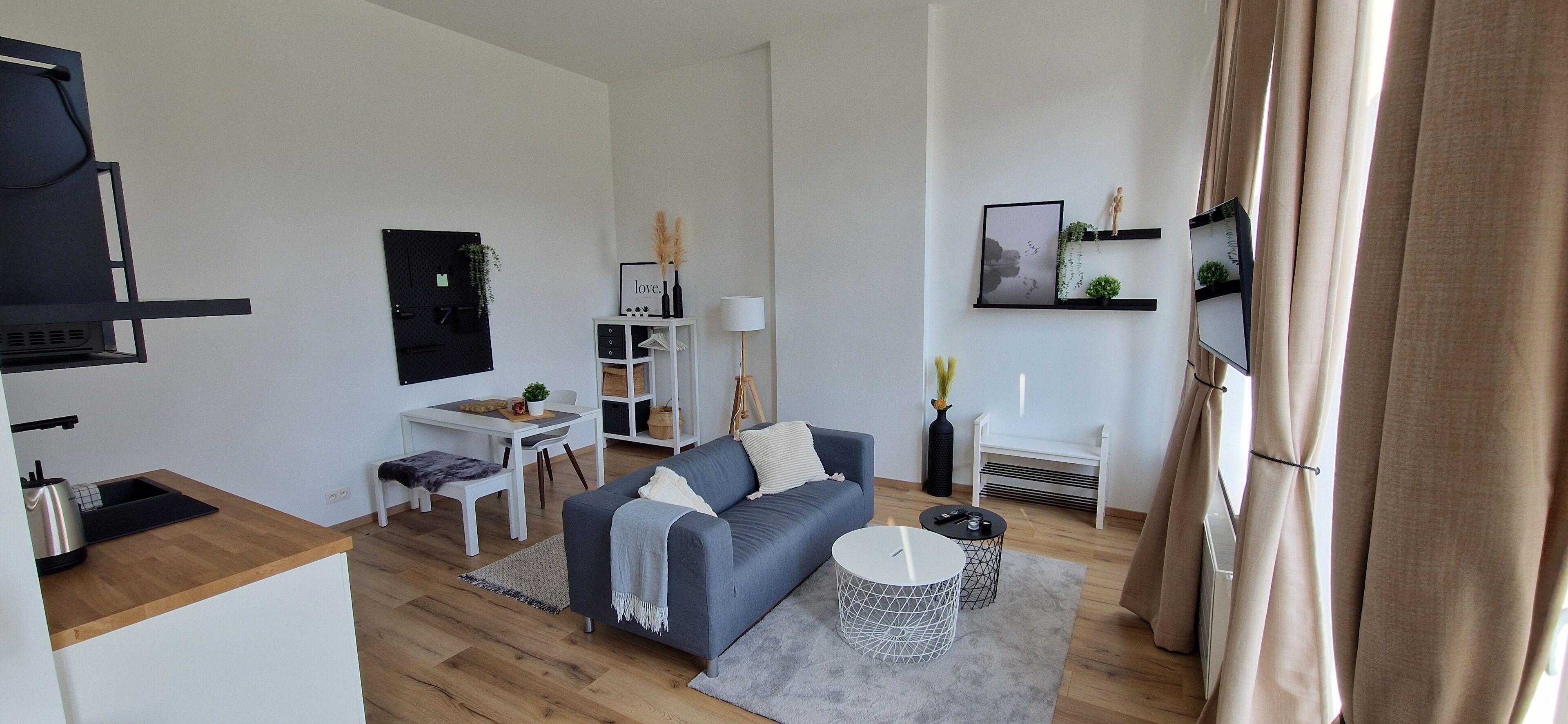 Una sala de estar moderna con un sofá gris, una mesita redonda, un televisor y una zona de cocina con estufa y fregadero.