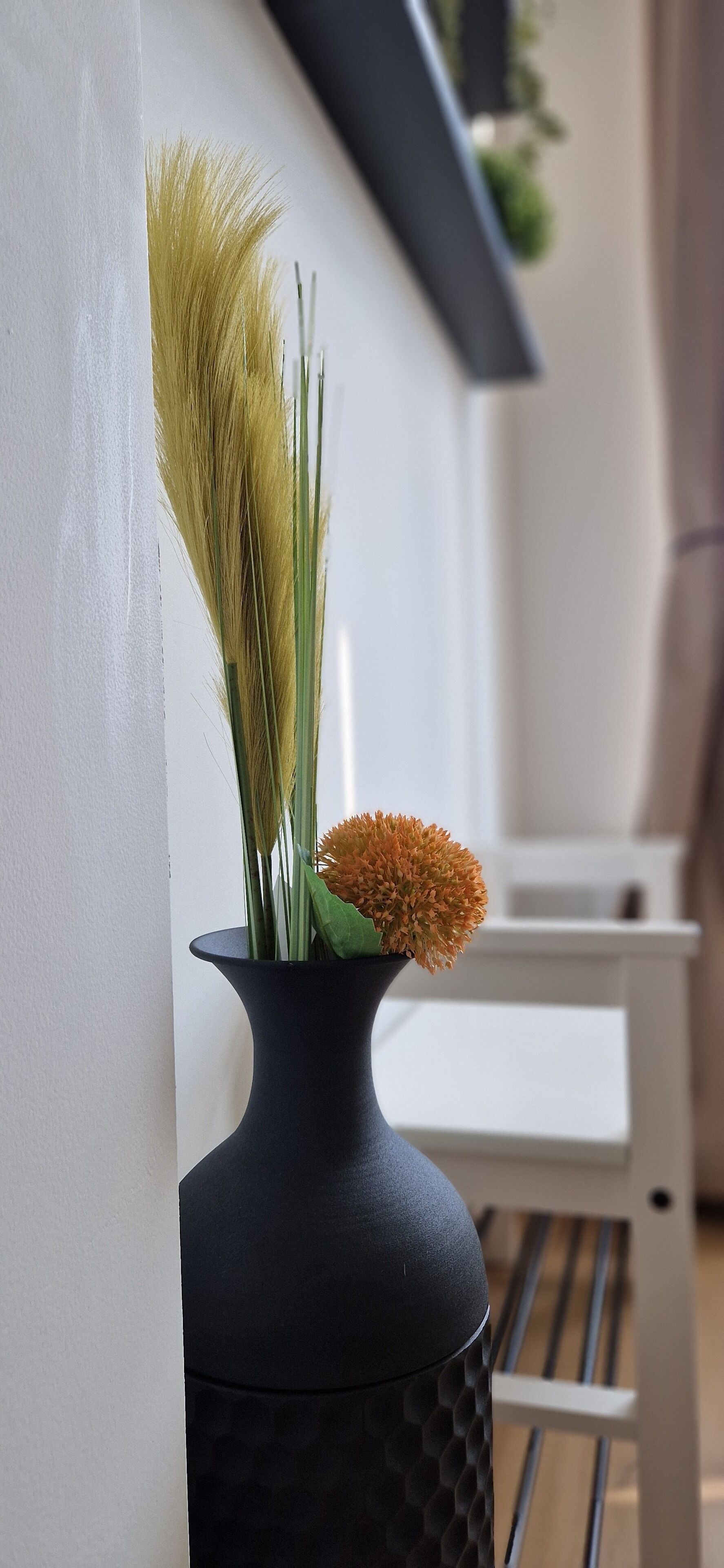 A black vase with dried plants and a single orange flower on a wooden floor.