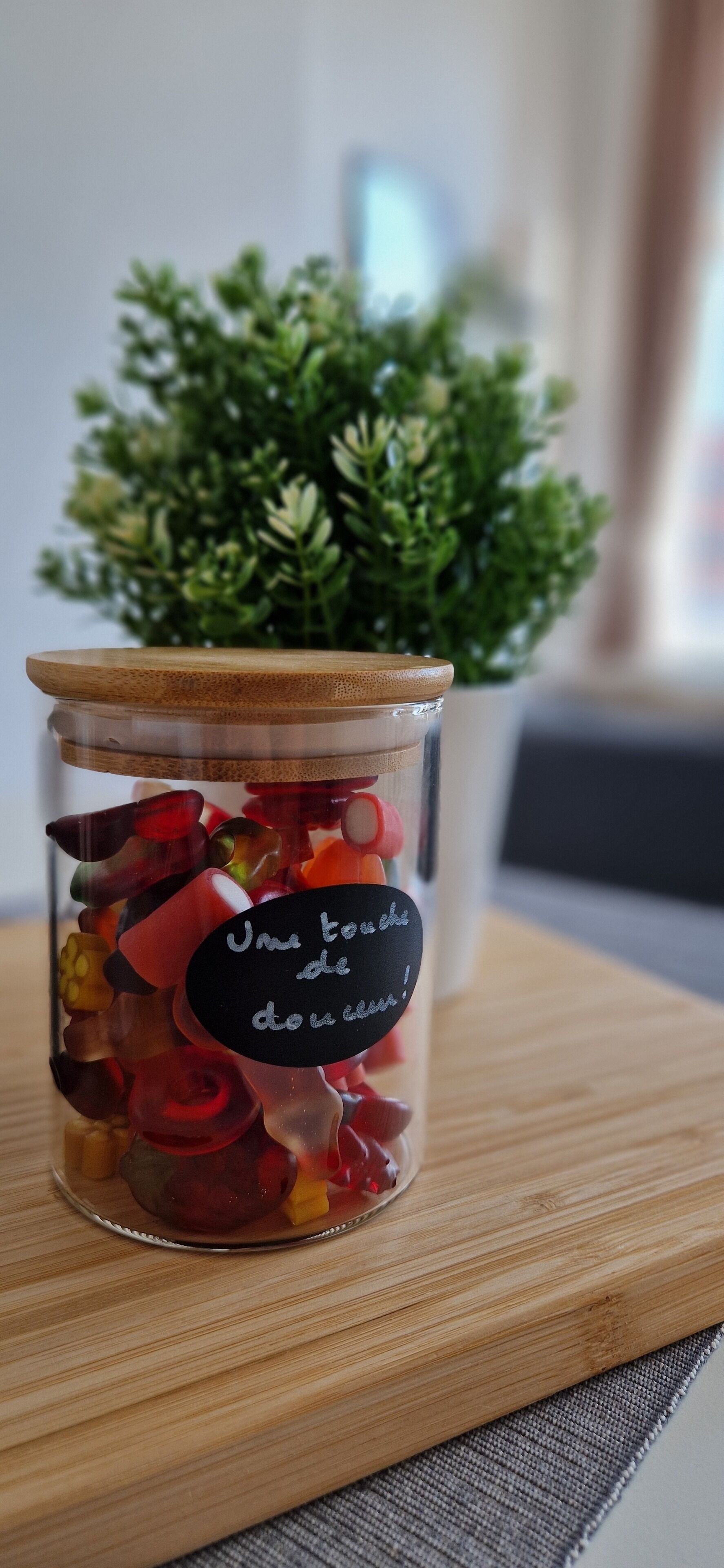 A glass jar with a wooden lid filled with colorful candies, placed on a wooden surface with a small potted plant in the background.