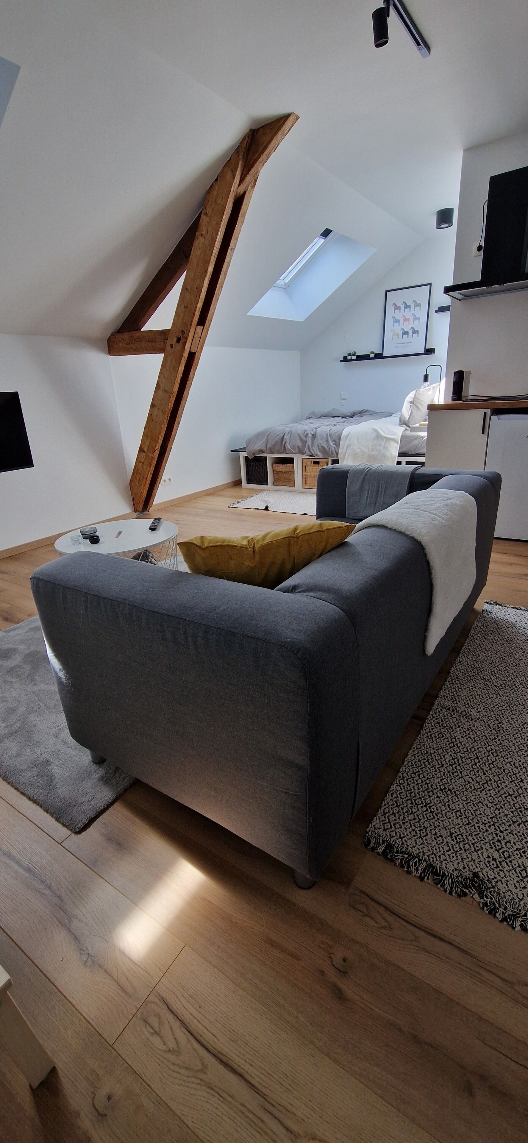 A modern loft bedroom with a sloped ceiling, a gray sofa, a small round table, and a bed with a white headboard.