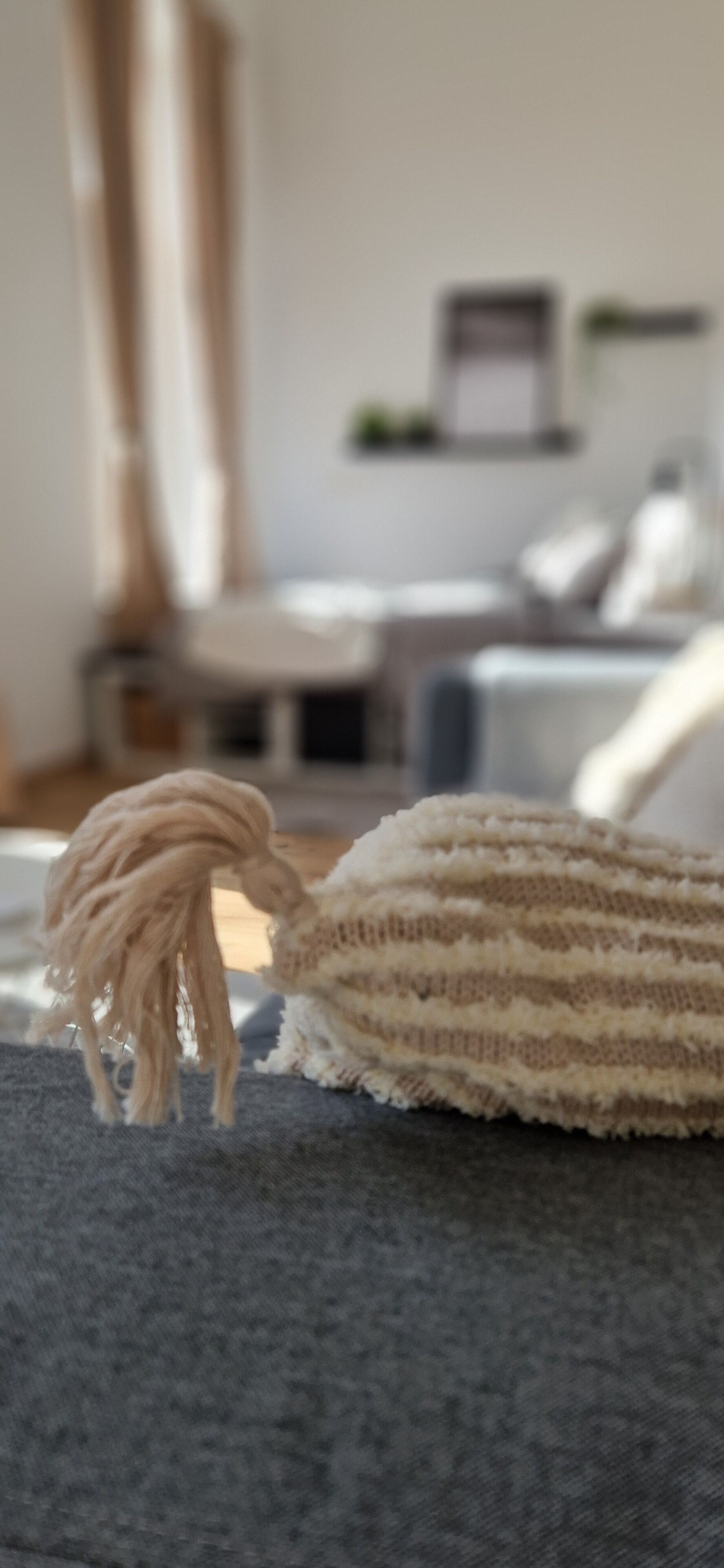 En närbild på ett stickat föremål med en tofs, placerat på en grå soffa.