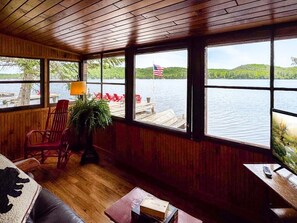 Une cabane aux murs en bois et aux grandes fenêtres donnant sur un lac, avec une chaise rouge, une lampe et une télécommande posées sur une table.