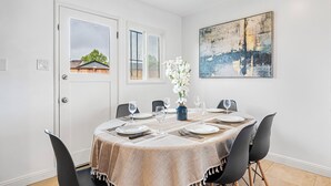 A dining area with a table set for six, a vase of white flowers, and a painting on the wall.