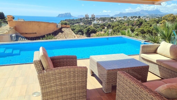 A poolside area with wicker furniture and a view of the sea and town. - Luxury villa with stunning sea views, walking distance from El Portet beach. (Teulada)