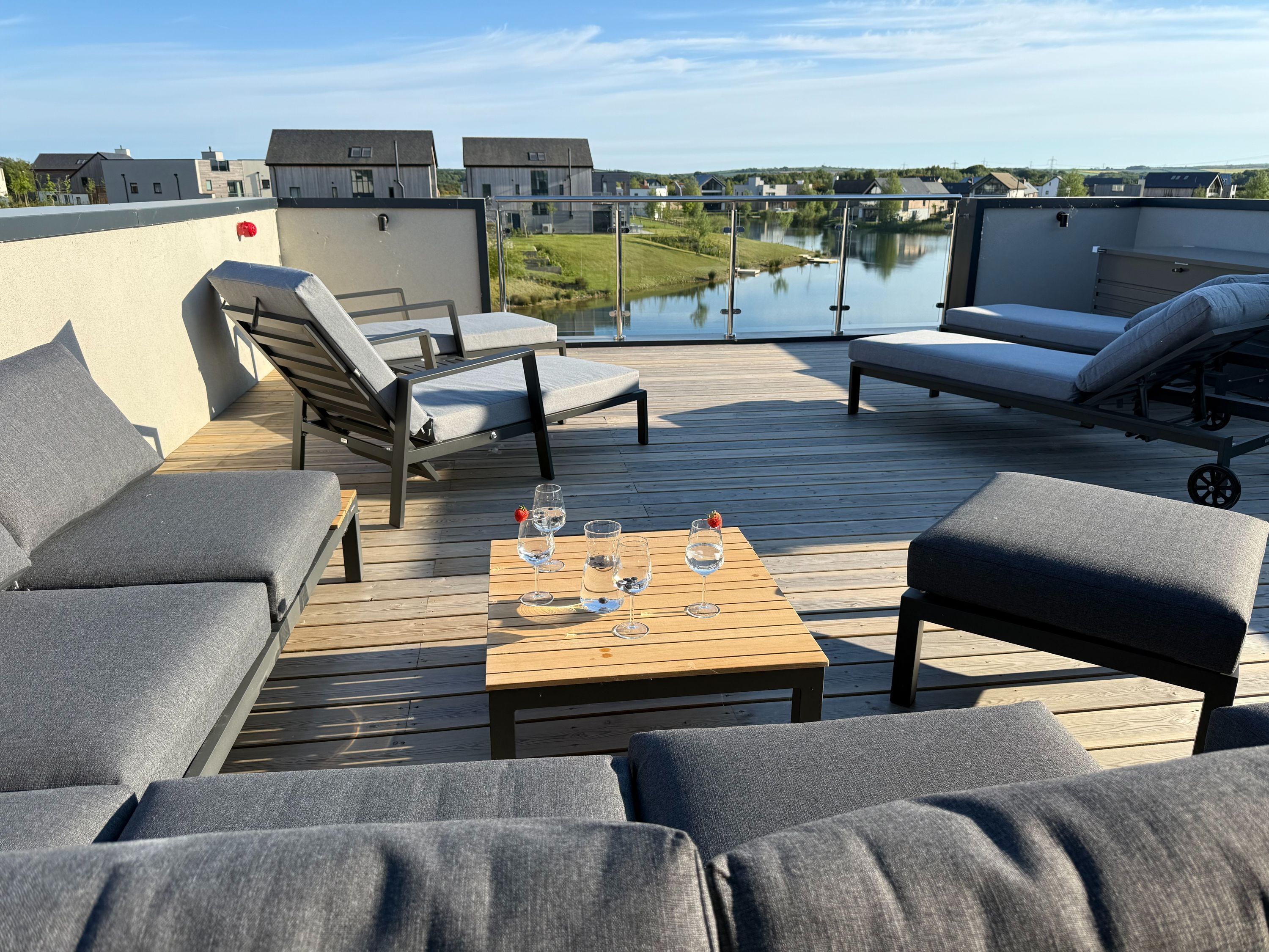 Una terraza con asientos acolchados de color gris, una mesa de madera con vasos y una botella, y una vista de un lago y edificios al fondo.