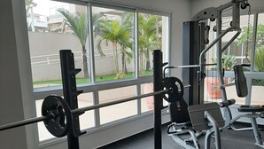 Ein Fitnessstudio mit Krafttrainingsgeräten, einer Bank und Blick auf Palmen durch große Fenster.