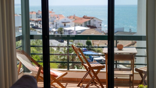 A balcony with a view of the sea and buildings. There are wooden chairs and a table with a bowl on it. - Apartamento Ouri o by Acasadascasas (Mafra)