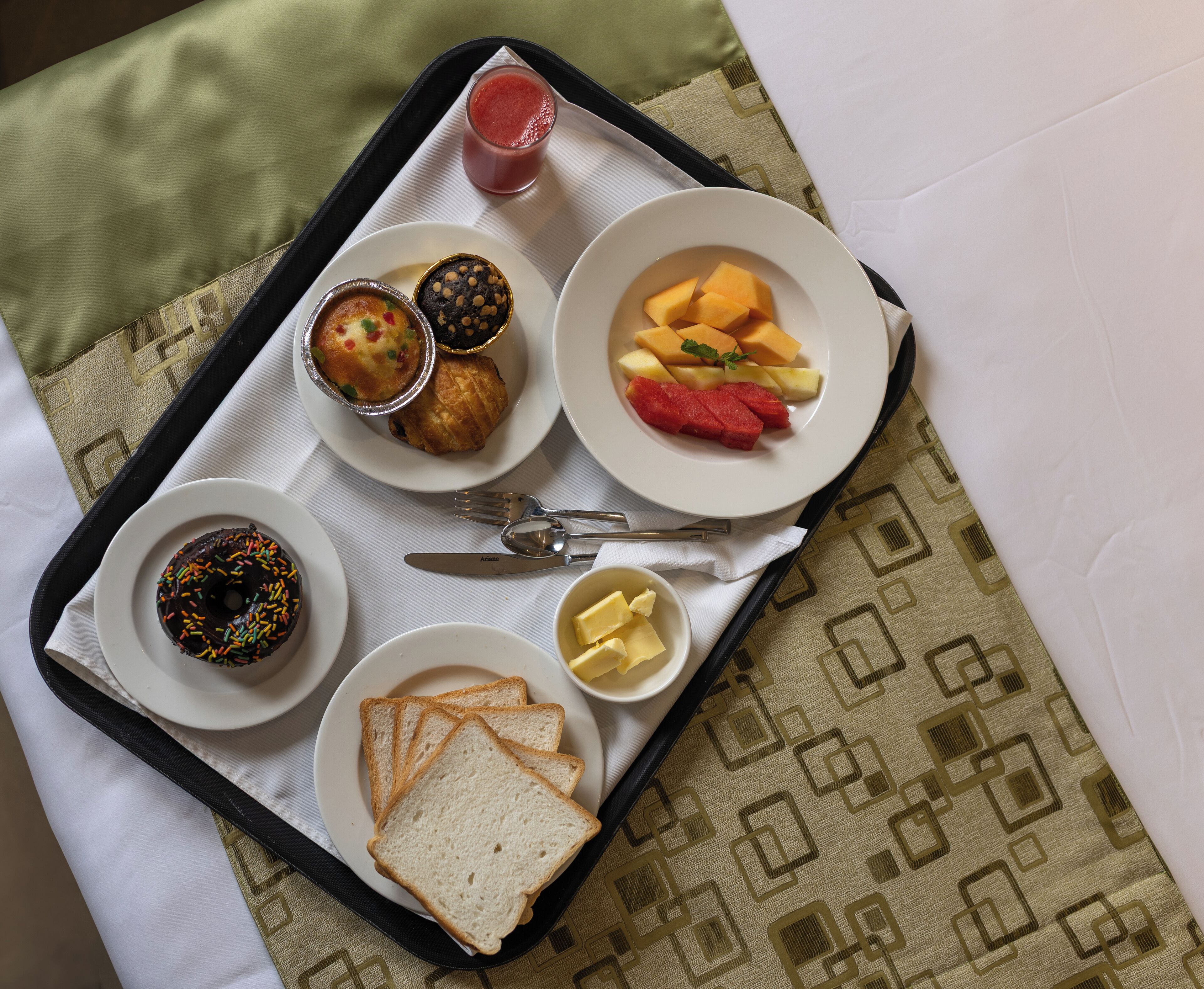 A tray with food items including bread, muffins, a fruit plate, and a glass of juice, placed on a table with a patterned tablecloth.