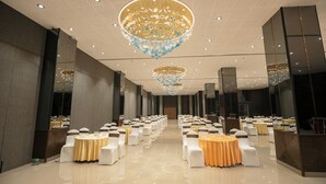 A spacious banquet hall with multiple round tables, white and orange tablecloths, and a large chandelier. - Biznotel by Pride Surat (Surat)