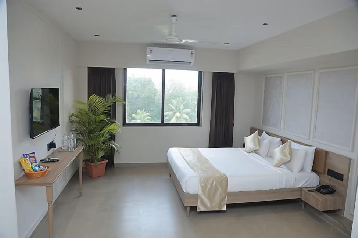 A hotel room with a bed, a TV, a desk with a plant, and a window with curtains.