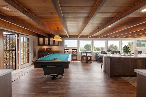 Une table de billard dans une pièce au plancher de bois, un coin repas avec des chaises et une cuisine avec un comptoir.
